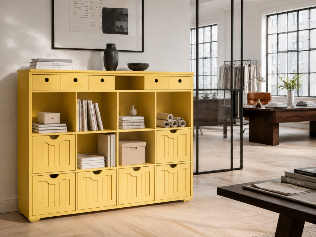Banana-yellow storage cabinet filled with books, boxes, fabric rolls and decorative objects in a bright urban design studio with pale parquet flooring, black-framed glass walls and dark wooden furniture.